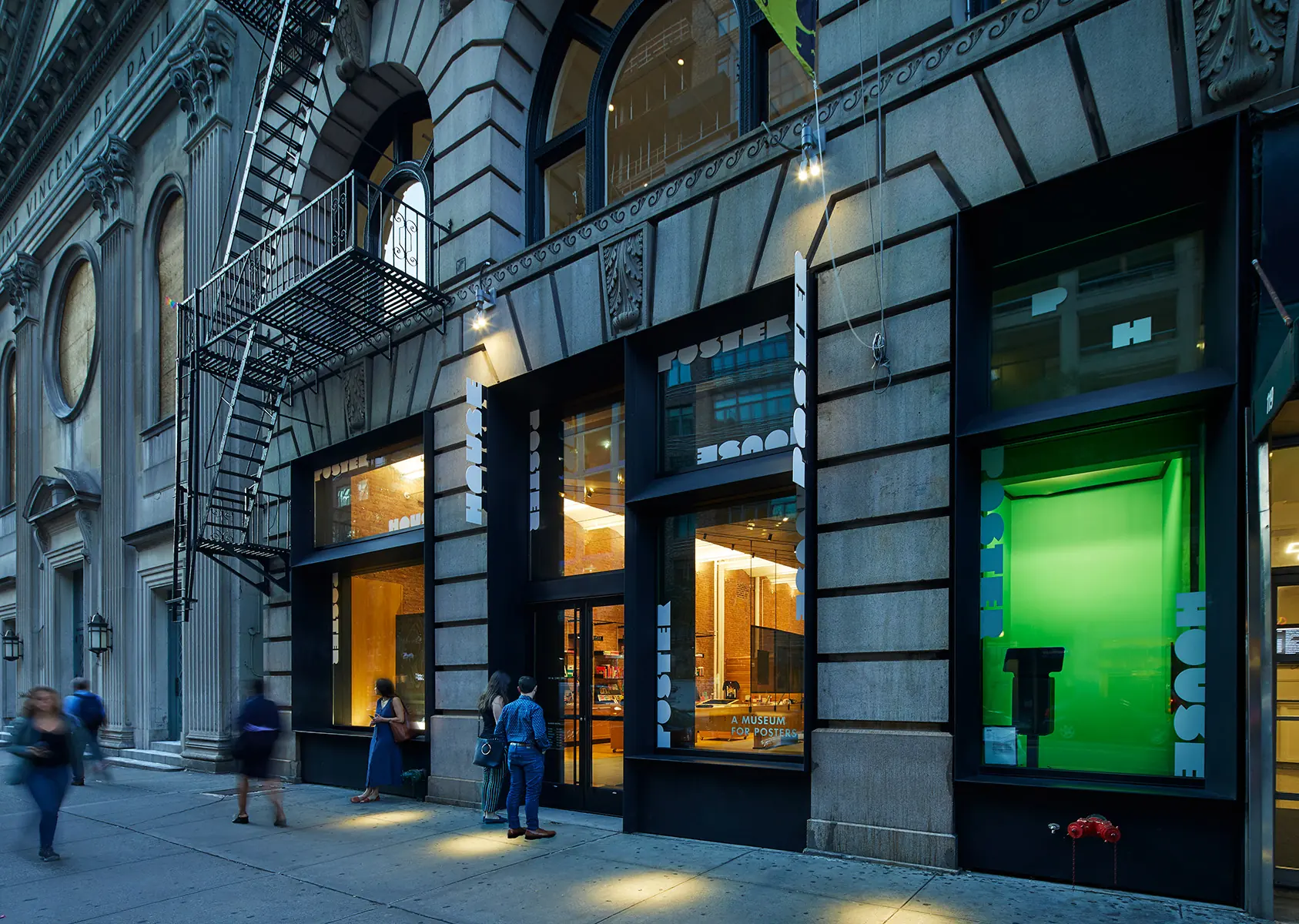 Street-level exterior of Poster House at dusk, with illuminated gallery windows and pedestrians.